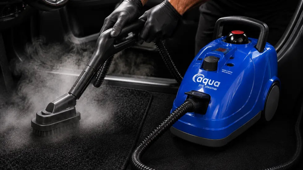 Close-up of the Aqua Pro Steamer being used to steam clean a vehicle floor mat, showing a professional auto detailing setup with visible vapor and handheld cleaning attachment