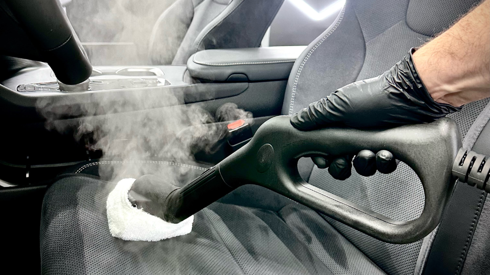 Person using a steam cleaner with a white cloth attachment to clean the seat of a car interior, with steam visibly rising from the device, demonstrating effective upholstery cleaning