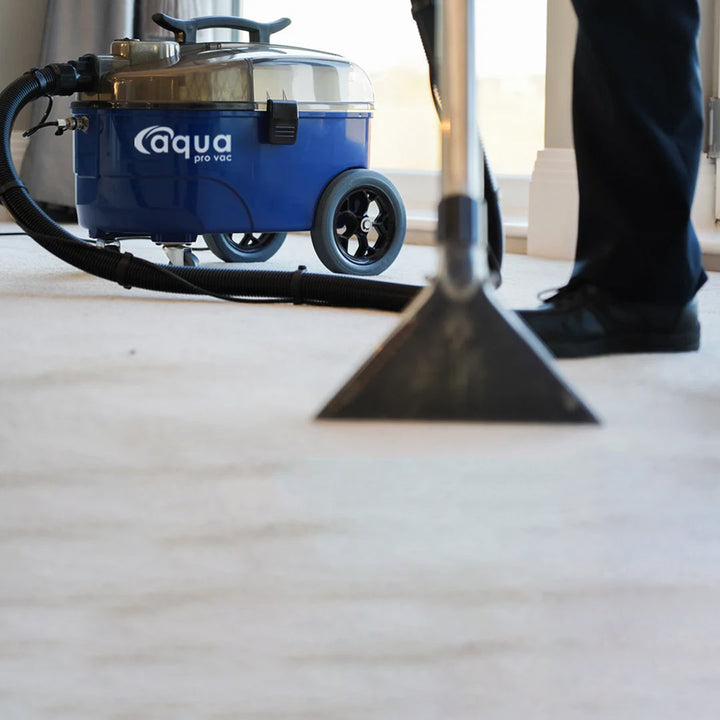 Aqua Pro Vac in use, showcasing the blue extractor machine and the carpet nozzle removing dirt from a white carpet.