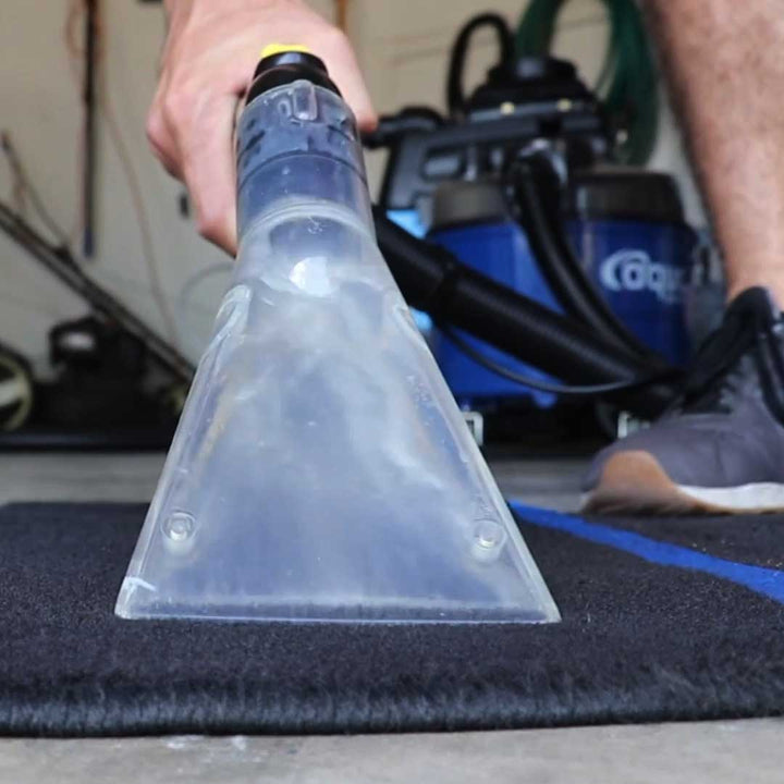 Close-up of the upholstery nozzle extracting dirt from a black vehicle mat, with the Aqua Pro Vac visible in the background.